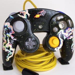 May include: A black video game controller with a glittery finish, featuring hand-painted colorful nebula clouds and a small rocket ship graphic. The controller includes a gray analog stick, a yellow C-stick, clear buttons, and a long yellow cord.