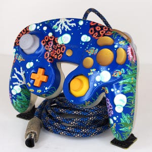 May include: A blue video game controller featuring a hand-painted underwater reef scene with coral, seaweed, and small fish. The controller includes a gray analog stick, a yellow C-stick, an orange D-pad, and a braided blue and black cable.