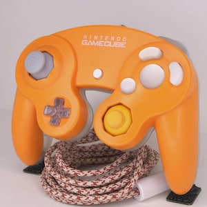 May include: Orange Nintendo GameCube controller with gray and white buttons, a gray joystick, and a yellow center button. The controller has the words "NINTENDO GAMECUBE" printed on it. A patterned cable is connected to the controller.