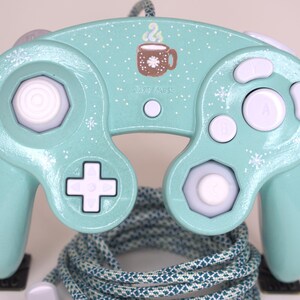 Puede incluir: Un mando de videojuegos de color verde menta claro con patrones de copos de nieve blancos y un gr&aacute;fico de una taza marr&oacute;n con vapor sobre el bot&oacute;n START/PAUSE. El mando tiene botones blancos, un stick anal&oacute;gico blanco, un stick C blanco y un cable trenzado azul verdoso.
