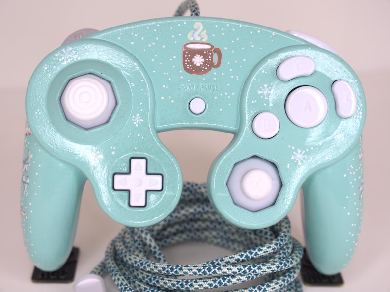 Puede incluir: Un mando de videojuegos de color verde menta claro con patrones de copos de nieve blancos y un gr&aacute;fico de una taza marr&oacute;n con vapor sobre el bot&oacute;n START/PAUSE. El mando tiene botones blancos, un stick anal&oacute;gico blanco, un stick C blanco y un cable trenzado azul verdoso.