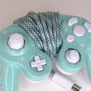 Puede incluir: Un mando de videojuegos de color verde menta con botones blancos, un joystick anal&oacute;gico blanco y una cruceta blanca. El mando est&aacute; decorado con peque&ntilde;os patrones de copos de nieve blancos y envuelto en un cable trenzado azul cerceta y blanco.
