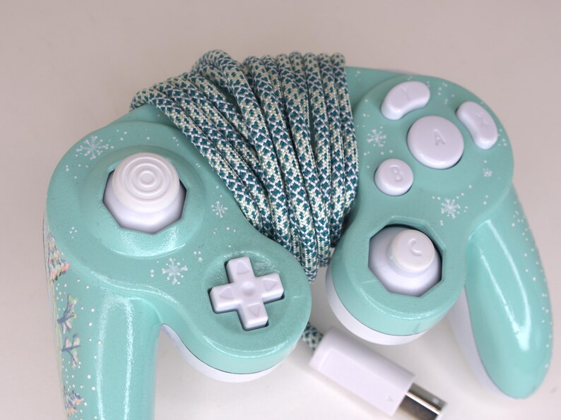Puede incluir: Un mando de videojuegos de color verde menta con botones blancos, un joystick anal&oacute;gico blanco y una cruceta blanca. El mando est&aacute; decorado con peque&ntilde;os patrones de copos de nieve blancos y envuelto en un cable trenzado azul cerceta y blanco.