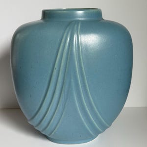 May include: A light blue ceramic vase with a rounded body and a narrow neck. The vase features three vertical, curved, raised lines on each side, creating a draped effect. The vase is a solid color and has a matte finish.