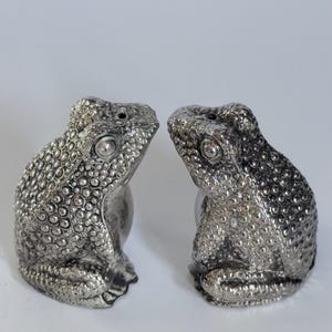 May include: A pair of silver-colored frog-shaped salt and pepper shakers. Each shaker has a textured surface with a detailed design, including eyes and small holes on top. The shakers are positioned facing each other.