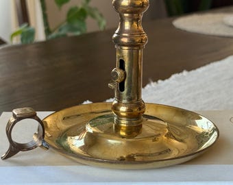 May include: A vintage brass candlestick holder with a round base and adjustable height mechanism. The candlestick has a classic design with a circular handle and is placed on a book titled "The Clockmaker's Daughter".
