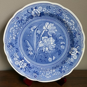 May include: Blue and white floral patterned plate with a scalloped edge. The plate features a central floral design and a border of smaller flowers and leaves. The plate is displayed on a wooden stand.