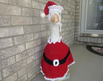 May include: A decorative goose statue wearing a crocheted Santa Claus outfit. The red outfit has a black belt with a white buckle, and the hat has a white trim and a white pom-pom. The goose is positioned in front of a brick wall.