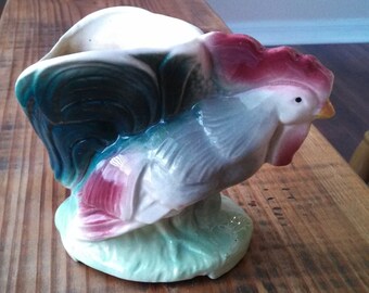 May include: A ceramic rooster planter with a green, blue, and pink glaze. The rooster is standing on a green base with a leaf design.