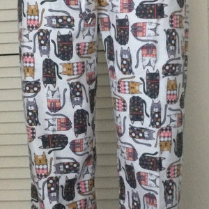 May include: White pajama pants with a repeating pattern of stylized cats in various colors, including black, gray, and tan. The pants have a drawstring waist.