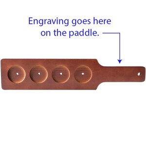 May include: A wooden paddle with four round indentations for holding drinks. The paddle has a brown stain and a smooth surface. The text "Engraving goes here on the paddle." is written above the paddle.