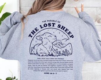 Può includere: Una felpa grigia con una grafica blu navy di un agnello e una mano. La grafica è circondata da un cerchio con il testo "The Parable of The Lost Sheep". Il testo "Then Jesus told them this parable" è sotto la grafica, seguito da un passaggio di Luca 15:3-7. Il testo "COLOR: SPORTS GREY" è nell'angolo in alto a destra dell'immagine.