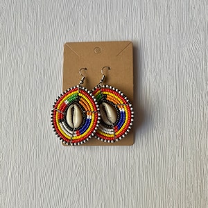 May include: A pair of colorful beaded earrings with a cowrie shell accent. The earrings are round and have a multi-colored design with red, yellow, blue, green, and black beads.