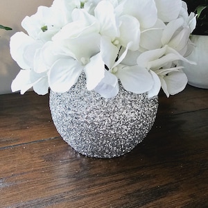 May include: A white hydrangea flower arrangement in a silver glitter vase. The vase is round and has a textured surface. The flowers are arranged in a cluster and have a natural, realistic look.