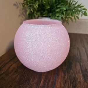 May include: A pink round vase with a glittery finish. The vase is made of ceramic and has a smooth, rounded shape.