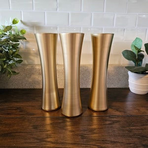 May include: Three gold-colored glass vases with a narrow base and a wide opening. The vases are arranged in a row on a wooden surface.