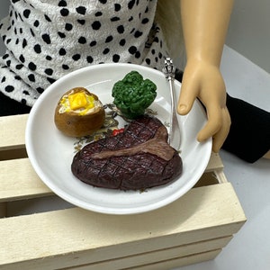 May include: A white plate with a toy steak, a toy baked potato with butter, and a toy broccoli floret. The plate is on a wooden crate.