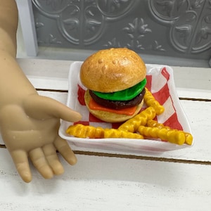 May include: A toy hamburger with lettuce, tomato, and cheese on a white plate with yellow french fries. The plate has a red and white checkered pattern.