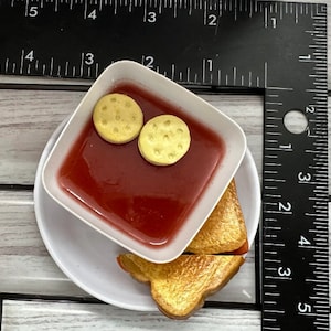 May include: A white plate with a white bowl filled with red soup and two yellow crackers. A grilled cheese sandwich is on the plate.