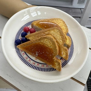 May include: A white plate with a blue and white floral design border. Three slices of French toast are arranged on the plate, topped with a sweet syrup. A few red and blue berries are scattered around the toast.
