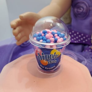 May include: A clear plastic cup filled with pink and blue candy sprinkles. The cup has a label that says "Dippin' Dots Ice Cream".