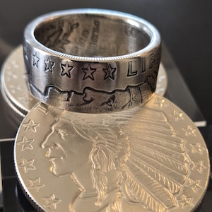 May include: A silver ring made from a modified US gold coin. The ring features the inscription "LIBERTY" and five stars. The ring is sitting on top of a silver coin with the profile of an indigenous person wearing a headdress.