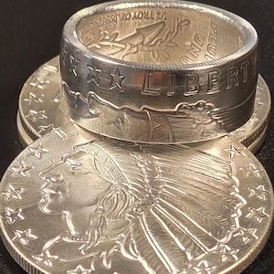 May include: A silver ring made from a US silver dollar coin. The ring features the words "LIBERTY" and "IN GOD WE TRUST" on the band. The ring is sitting on top of two silver dollar coins, one with the image of an American Indian chief and the other with the image of an eagle.