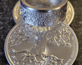 May include: A silver ring with an engraved tree design, resting on a silver coin with a tree of life motif. The ring is marked with the year 2025. The coin has intricate details, and the overall aesthetic is metallic and detailed.