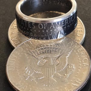 Könnte beinhalten: Ein Silberring in Form einer halben Dollar-Münze. Der Ring ist aus einer halben Dollar-Münze gefertigt, die geschnitten und zu einem Ring geformt wurde. Der Ring ist schwarz und trägt die Worte "United States of America" auf der Band. Der Ring liegt auf zwei weiteren halben Dollar-Münzen.
