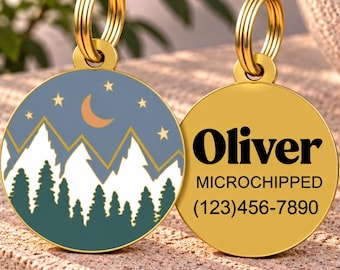 May include: Two round gold-toned metal pet ID tags with split rings. One tag features a graphic of white mountains, dark green pine trees, a crescent moon, and stars against a blue sky. The other tag displays the text Oliver, MICROCHIPPED, and (123)456-7890.