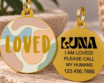 Op de afbeelding: Twee ronde huisdierpenningen van goudkleurig metaal met ringen. Eén penning heeft een golvend, pastelkleurig patroon met het woord LOVED in goud. De andere is effen goudkleurig met zwarte tekst: LUNA, I AM LOVED! PLEASE CALL MY HUMANS 123.456.7890.