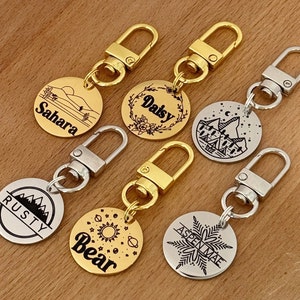 May include: Six gold and silver keychains with round metal tags. Each tag has a different design and name engraved on it. The names are Sahara, Daisy, Aspen Mae, Rusty, Bear, and Granite.