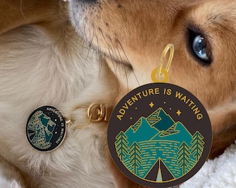 Pode incluir: Uma medalha redonda para cães com o texto "ADVENTURE IS WAITING" e um design de montanha e floresta turquesa. A medalha está presa a um fecho dourado. A pelagem do cão é castanho-claro e branca.