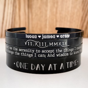 May include: Four black metal bracelets stacked on top of each other. The top bracelet has the names "lucas james crew" engraved on it. The second bracelet has the date "VII.XIII.MMXIX" engraved on it. The third bracelet has the text "Grant me the serenity to accept the things I cannot change; the things I can; And wisdom to know the difference." engraved on it. The bottom bracelet has the text "ONE DAY AT A TIME." engraved on it.