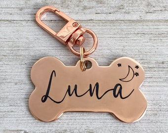 May include: Rose gold bone-shaped dog tag with a black engraved inscription that reads "Luna" and a crescent moon with stars design.