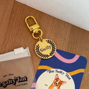 Könnte beinhalten: Ein goldenes Hundehalsband mit einem Kranz-Design und dem eingravierten Namen "Sicily". Das Halsband ist an einem goldenen Splitt-Ring befestigt. Das Halsband liegt neben einer Visitenkarte mit einem Cartoon-Hund und dem Text "The Salty Hut Custom Pet Tags".