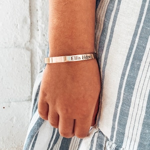 May include: A gold-toned bracelet with the name "Ellis Hope" engraved on it. The bracelet is worn on a person's wrist.