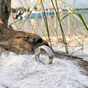 May include: A silver wedding band with an inscription on the inside of the band. The ring is lying on a white sandy surface with a piece of driftwood in the background.
