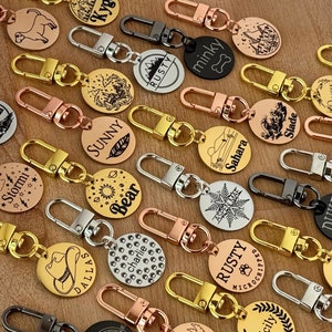 May include: A collection of personalized dog tags with various designs and engravings. The tags are made of metal and come in different colors, including gold, silver, and black. Some of the tags feature names, while others have images of animals, mountains, and other designs.