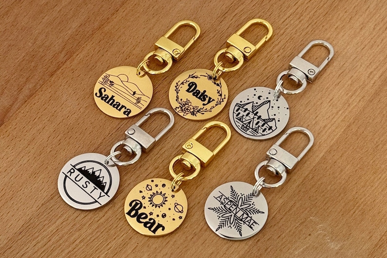 May include: Six gold and silver keychains with round metal tags. Each tag has a different design and name engraved on it. The names are Sahara, Daisy, Aspen Mae, Rusty, Bear, and Granite.