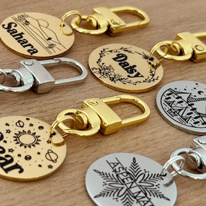 May include: Five gold and silver round pet ID tags with black engraved text and designs. The tags feature the names "Sahara", "Daisy", "Star", "Aspenmae", and a camping scene. Each tag is attached to a gold or silver metal split ring.