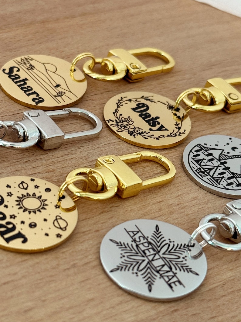 May include: Five gold and silver round pet ID tags with black engraved text and designs. The tags feature the names "Sahara", "Daisy", "Star", "Aspenmae", and a camping scene. Each tag is attached to a gold or silver metal split ring.