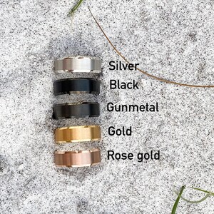 May include: Five different colored rings stacked on top of each other on a sandy surface. The rings are silver, black, gunmetal, gold, and rose gold.