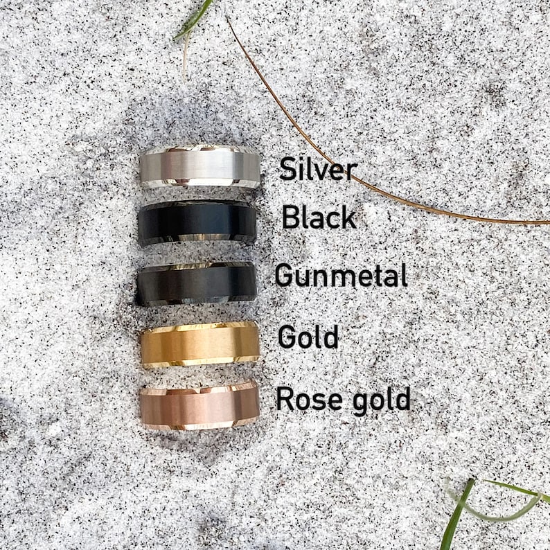 May include: Five different colored rings stacked on top of each other on a sandy surface. The rings are silver, black, gunmetal, gold, and rose gold.