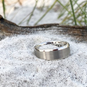 May include: A brushed silver wedding band with a beveled edge. The ring is sitting on a bed of white sand.