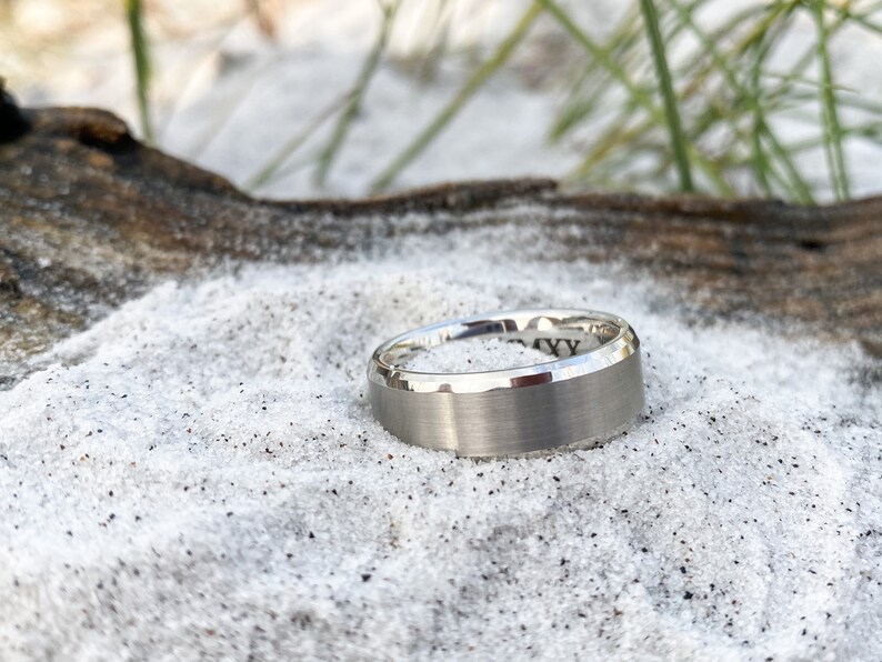 May include: A brushed silver wedding band with a beveled edge. The ring is sitting on a bed of white sand.