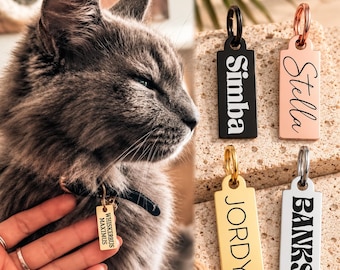 May include: Four rectangular metal pet ID tags with rounded top edges and split rings. The tags feature engraved names: black with 'Simba', rose gold with 'Stella', gold with 'JORDY', and silver with 'BANKS'. One tag reads 'WHISKERBUS MAXIMUS'.