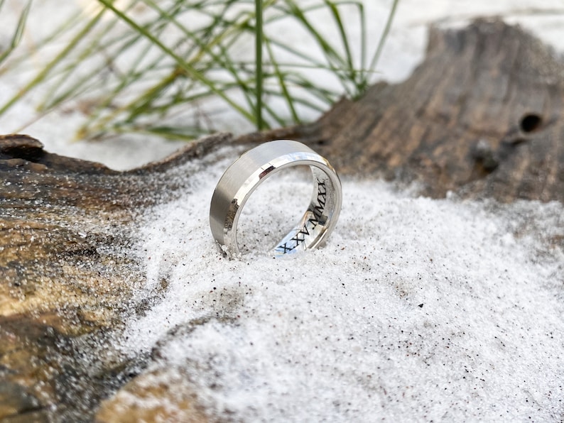May include: A silver wedding band with a beveled edge and a date inscription on the inside of the band. The ring is resting on a piece of driftwood and sand.