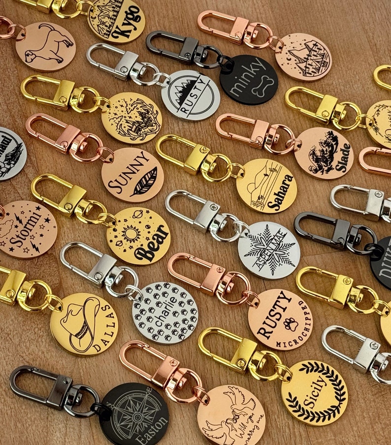 May include: A collection of personalized dog tags with various designs and engravings. The tags are made of metal and come in different colors, including gold, silver, and black. Some of the tags feature names, while others have images of animals, mountains, and other designs.