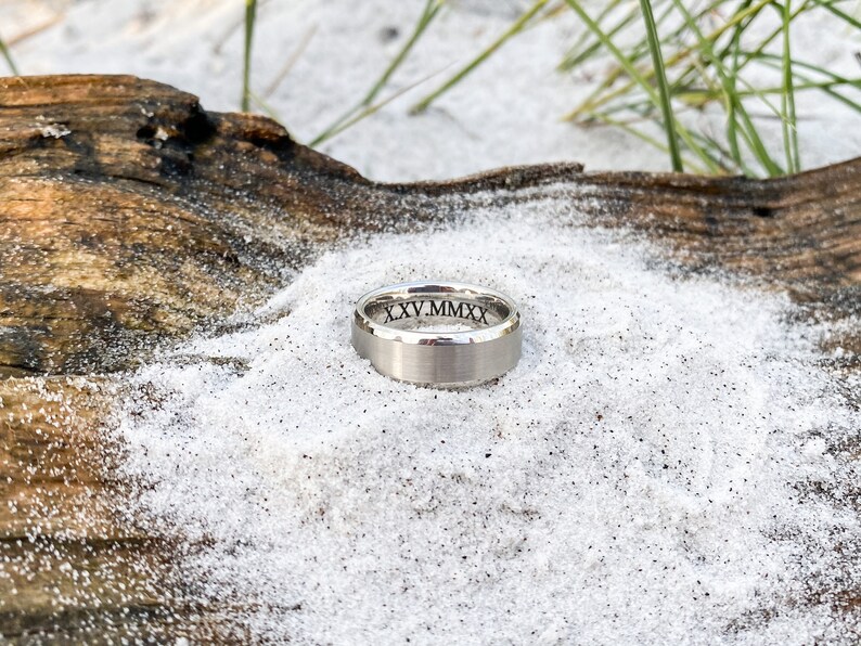 May include: A silver wedding band with a brushed finish and the inscription "XXV.MMXX" on the inside of the band.
